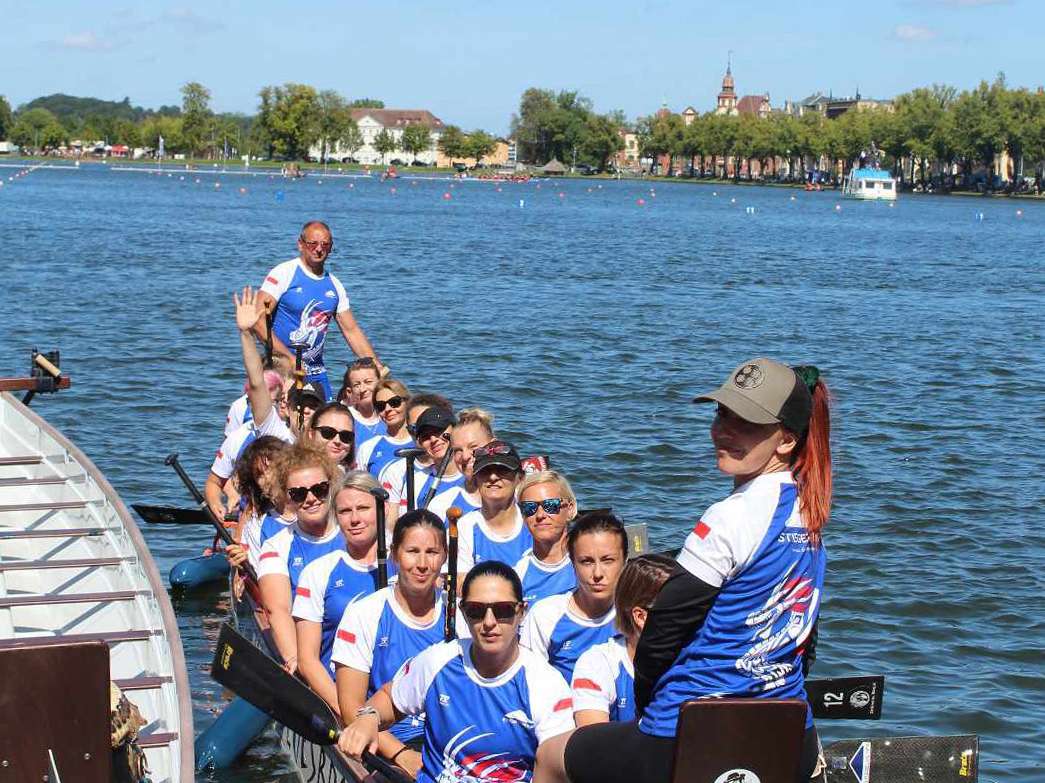 The dragon boat team from Stiegelmeyer Stolno sits in its boat, next to another empty boat, on the Pfaffenteich in Schwerin and smiles at the camera.