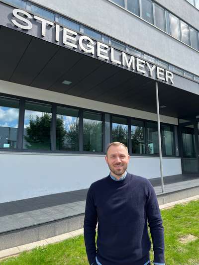 Anton Schlee steht auf dem Bürgersteig vor dem Eingang zum Stiegelmeyer-Verwaltungsgebäude in Herford und lächelt in die Kamera.