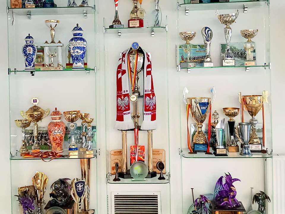A large number of trophies in different shapes and colours stand on a glass shelf in front of a white wall.