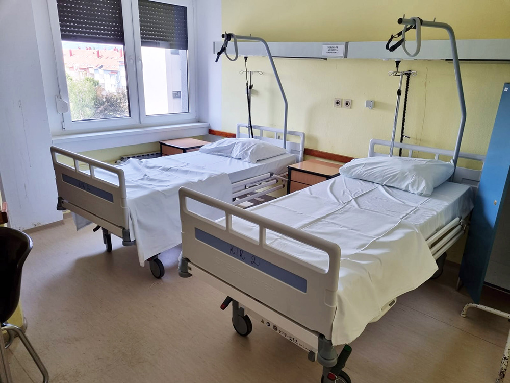 Two hospital beds stand in a room in front of a light yellow wall